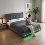 Woman sitting on a custom grey velvet LED bed frame, demonstrating premium B2B bedroom furniture quality by MMK Furniture Makers USA.